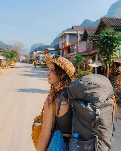 Eine junge Frau trägt einen Rucksack auf dem Rücken. Sie geht eine Straße in Südostasien entlang und schaut sich um.