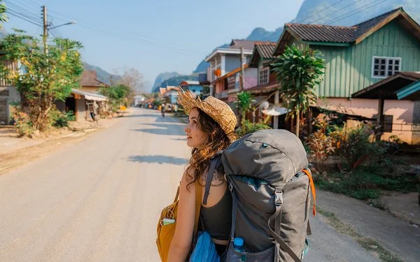 Eine junge Frau trägt einen Rucksack auf dem Rücken. Sie geht eine Straße in Südostasien entlang und schaut sich um.