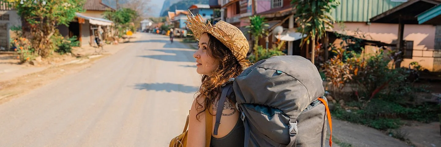 Eine junge Frau trägt einen Rucksack auf dem Rücken. Sie geht eine Straße in Südostasien entlang und schaut sich um.
