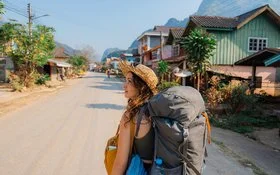 Eine junge Frau trägt einen Rucksack auf dem Rücken. Sie geht eine Straße in Südostasien entlang und schaut sich um.