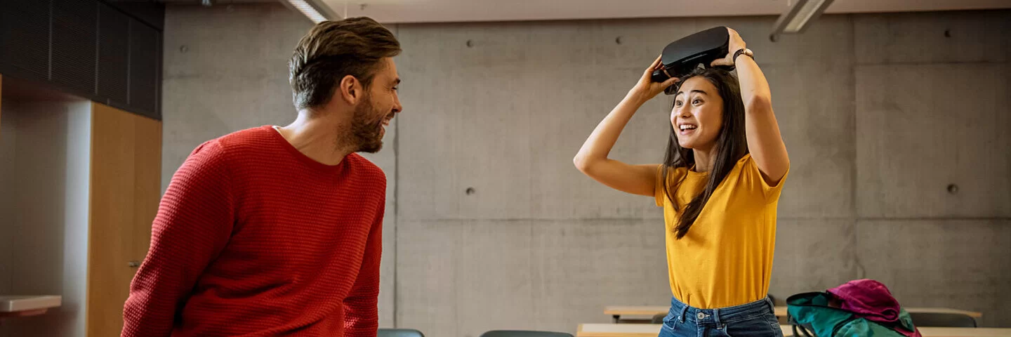 Eine junge Frau nimmt erstaunt lachend eine VR-Brille von ihrem Kopf. Sie schaut dabei zu einem ebenso lachenden Mann herüber.