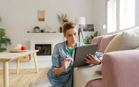 Junge Frau sitzt im Wohnzimmer auf dem Fußboden, ans Sofa gelehnt, und schaut konzentriert auf ein Tablet in ihrer Hand.