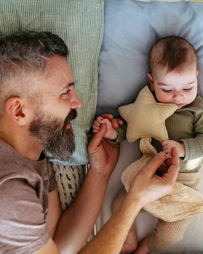 Ein Vater liegt neben seinem Baby im Gitterbett, blickt es zärtlich an und hält es an den Händen.
