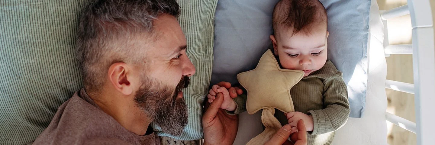 Ein Vater liegt neben seinem Baby im Gitterbett, blickt es zärtlich an und hält es an den Händen.