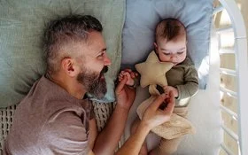 Ein Vater liegt neben seinem Baby im Gitterbett, blickt es zärtlich an und hält es an den Händen.
