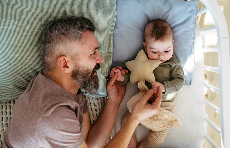 Ein Vater liegt neben seinem Baby im Gitterbett, blickt es zärtlich an und hält es an den Händen.