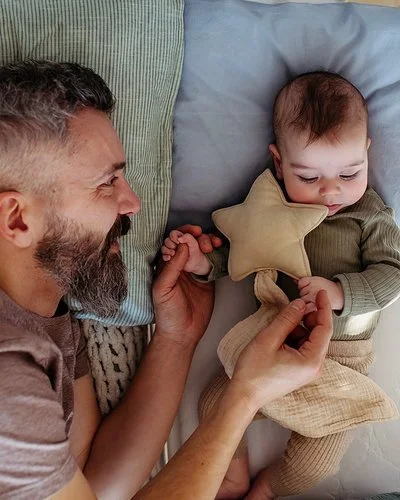 Ein Vater liegt neben seinem Baby im Gitterbett, blickt es zärtlich an und hält es an den Händen.