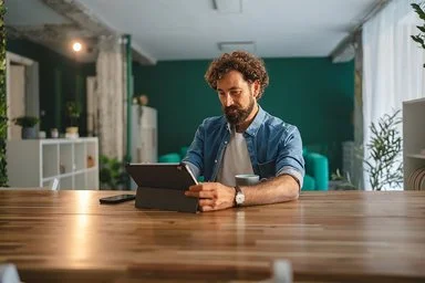 Eine Person mit lockigen Haaren sitzt an einem langen Holztisch und hält ein Tablet in beiden Händen. Die Person trägt ein hellblaues Hemd über einem weißen T-Shirt. Im Hintergrund sind Regale mit Pflanzen und Dekorationsgegenständen sowie eine grüne Wand zu sehen. Auf dem Tisch steht eine Tasse.
