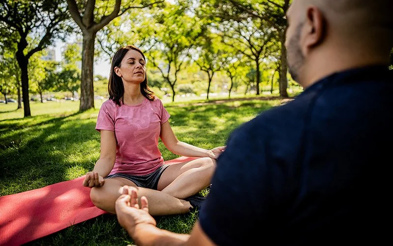 Eine Frau und ein Mann in mittlerem Alter in Sporkleidung meditieren auf Matten im Park.