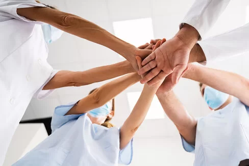 Vier Menschen in Krankenhauskleidung stehen im Kreis zusammen und legen die Hände übereinander. Das Ärzteteam der AOK-Zahnklinik lebt die Gemeinschaft.
