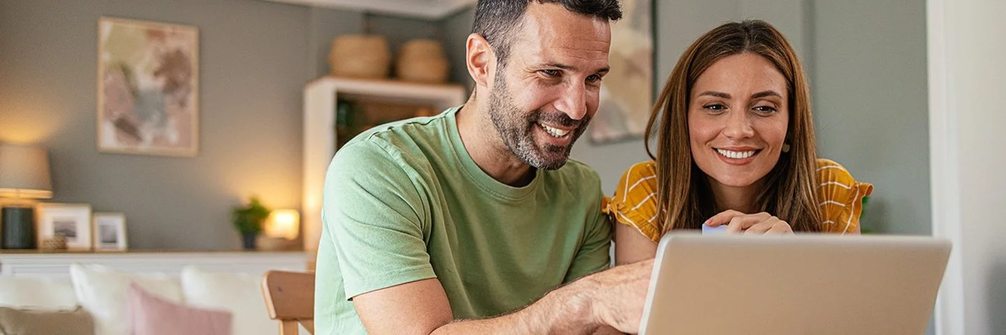 Zwei Personen sitzen an einem Holztisch und schauen gemeinsam auf einen geöffneten Laptop.