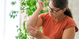 Eine Frau in einer Wohnung mit Grünpflanzen im Hintergrund. Sie blickt auf ihren rechten Ellenbogen und reibt sich die Haut mit einer weißen Creme ein.