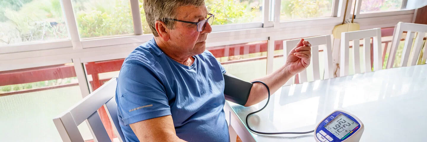Ein Mann mit Übergewicht sitzt an einem Tisch und misst seinen Blutdruck.