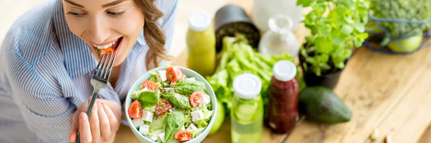 Eine junge Frau sitzt an einem Tisch und isst einen Salat mit Tomaten und Feta aus einer Schüssel.