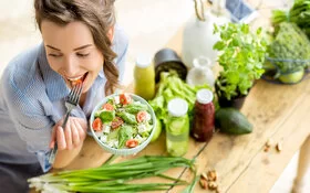 Eine junge Frau sitzt an einem Tisch und isst einen Salat mit Tomaten und Feta aus einer Schüssel.