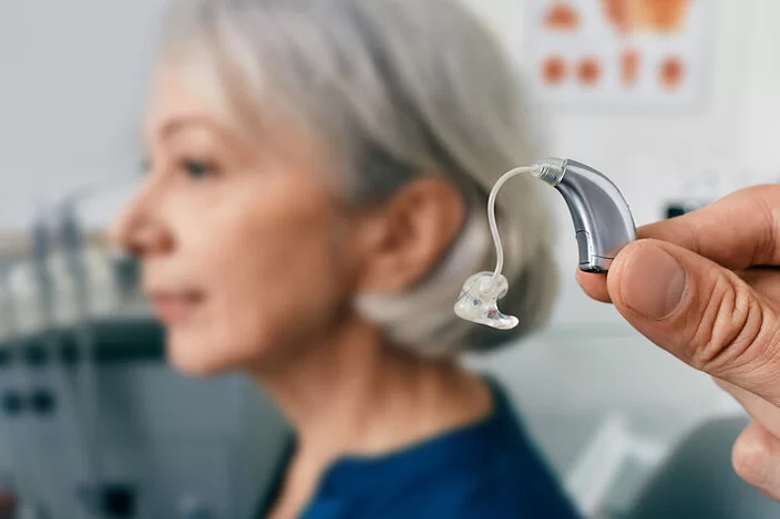 Person hält ein Hörgerät in die Kamera, im Hintergrund ist eine ältere Frau zu erkennen.