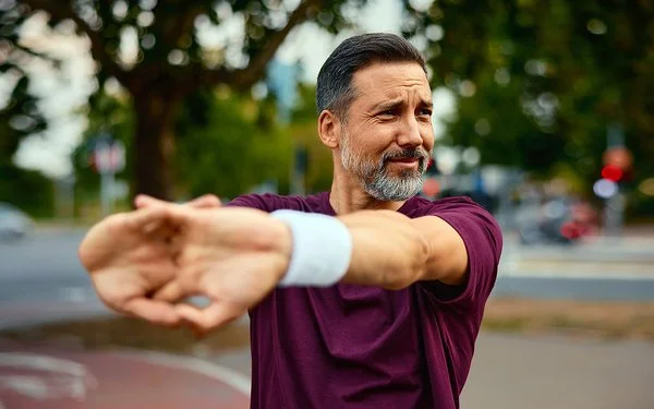 Ein Mann in dunkelrotem Sport-Shirt steht im Freien und dehnt seine Arme.