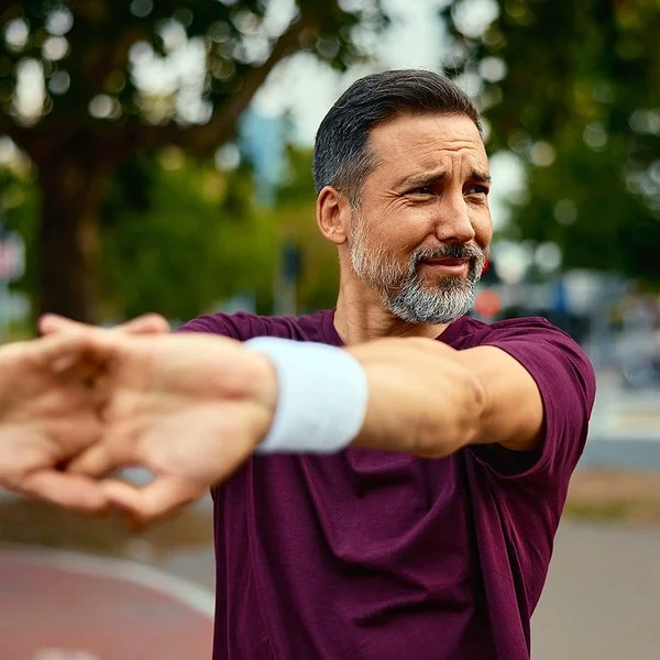 Ein Mann in dunkelrotem Sport-Shirt steht im Freien und dehnt seine Arme.