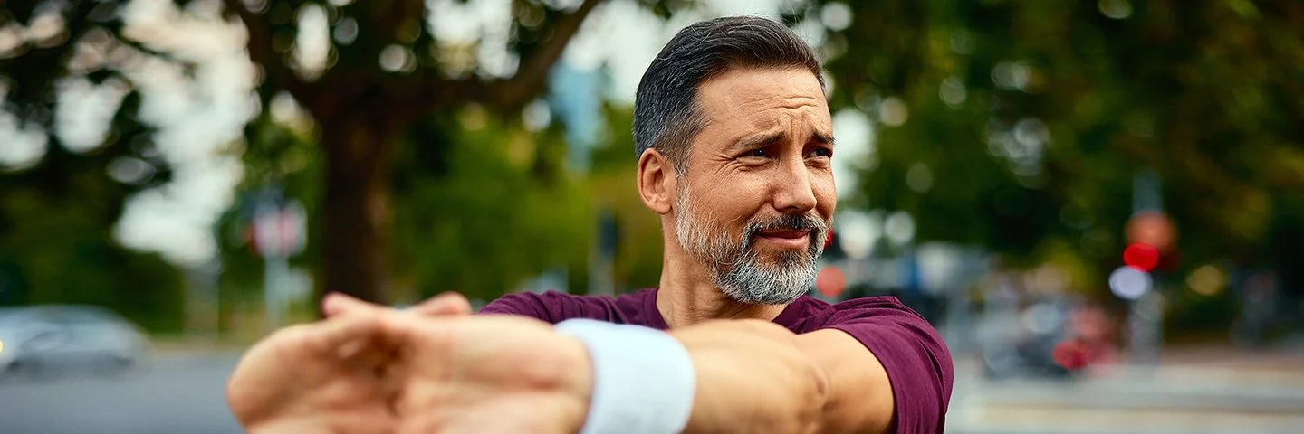 Ein Mann in dunkelrotem Sport-Shirt steht im Freien und dehnt seine Arme.