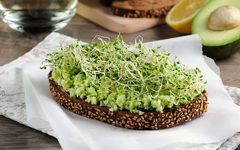 Ein Scheibe Graubrot mit Avocado-Frischkäse-Aufstrich und Kresse liegt auf einem Tisch neben halbierten Früchten.