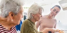 Zwei ältere und eine junge Frau stehen zusammen an einem Kochherd in einer Küche. Sie kochen gemeinsam und unterhalten sich. Auf der Arbeitsfläche liegen eine Tomate, ein Apfel, eine halbe Möhre und eine Ananas. Eine der beiden älteren Frauen schiebt in Scheiben geschnittene Möhren von einem Schneidebrett in einen Topf.