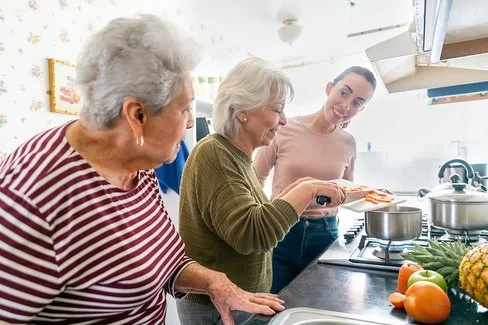Zwei ältere und eine junge Frau stehen zusammen an einem Kochherd in einer Küche. Sie kochen gemeinsam und unterhalten sich. Auf der Arbeitsfläche liegen eine Tomate, ein Apfel, eine halbe Möhre und eine Ananas. Eine der beiden älteren Frauen schiebt in Scheiben geschnittene Möhren von einem Schneidebrett in einen Topf.