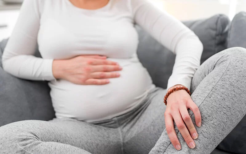 Eine Schwangere sitzt auf der Couch und hält sich den Bauch und das Bein.