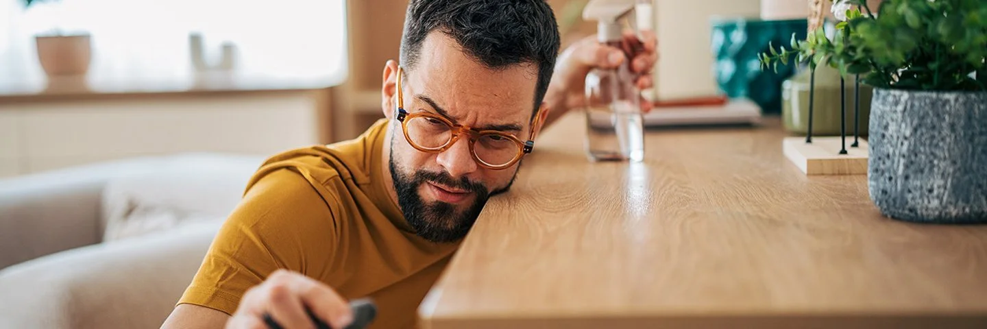 Ein mittelalter Man reinigt akkurat und mit kritischem Blick einen Tisch mit Tuch und Spray.