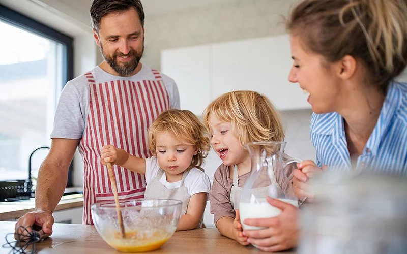 Vater, Mutter und zwei Kinder im Kindergartenalter rühren einen Kuchenteig in der Küche. Alle lachen und haben Spaß.