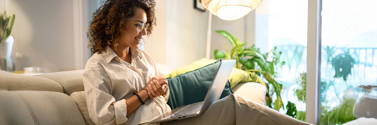 Eine junge Frau mit braunen, lockigen Haaren, Brille, weiß gestreiftem Hemd und beiger Hose sitzt lächelnd auf einem grauen Sofa. Ein Laptop liegt auf ihrem Schoß, ihre Hände sind ineinandergelegt. Im Hintergrund sind eine Pflanze und eine Stehlampe zu sehen.