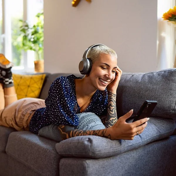 Eine junge Frau hat Kopfhörer auf und schaut auf dem Sofa liegend auf ihr Handy.