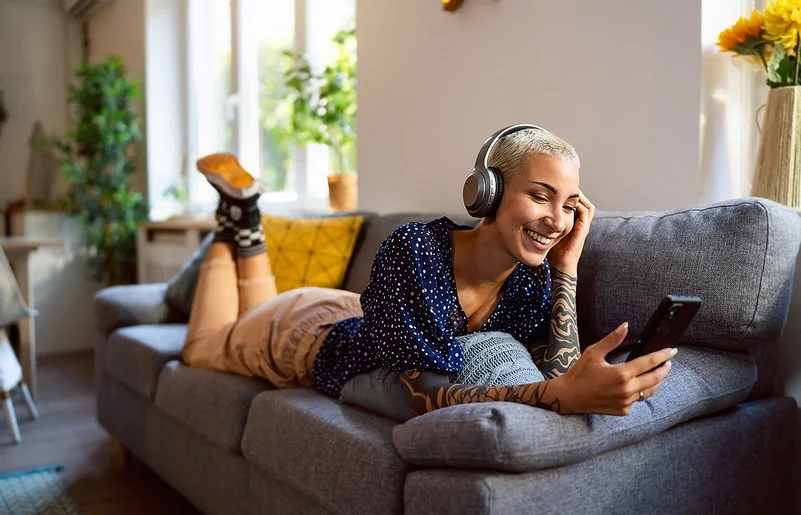 Eine junge Frau hat Kopfhörer auf und schaut auf dem Sofa liegend auf ihr Handy.