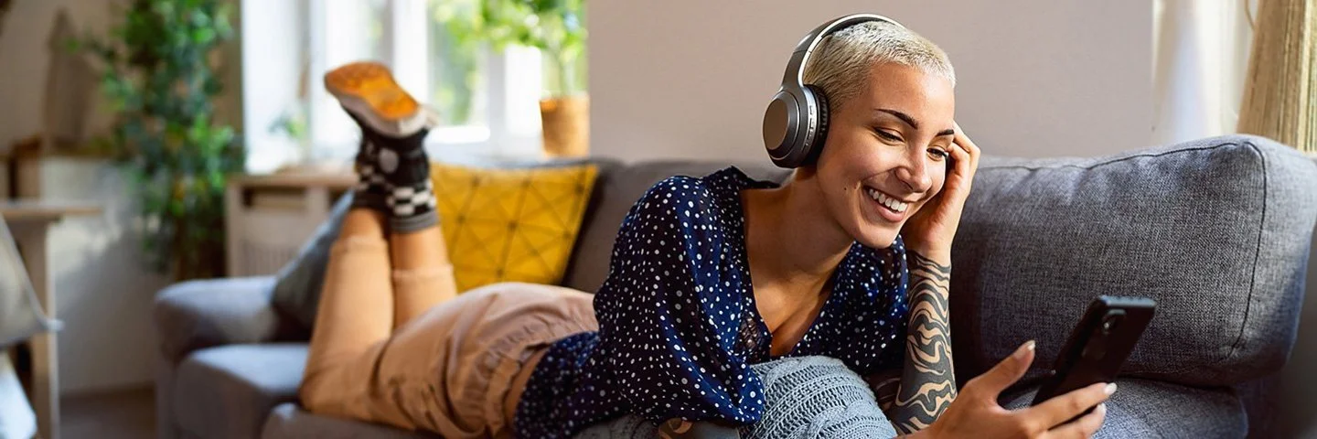 Eine junge Frau hat Kopfhörer auf und schaut auf dem Sofa liegend auf ihr Handy.