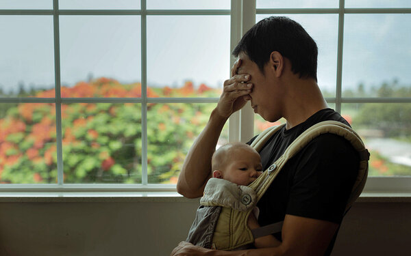 Ein Vater mit Baby vor der Brust reibt sich müde die Augen – er leidet unter Schlafmangel.