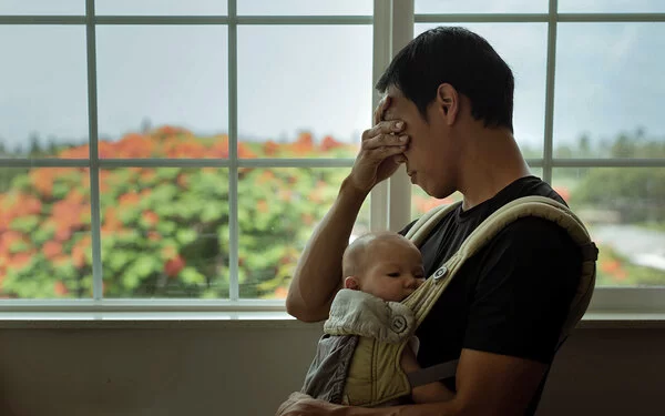 Ein Vater mit Baby vor der Brust reibt sich müde die Augen – er leidet unter Schlafmangel.