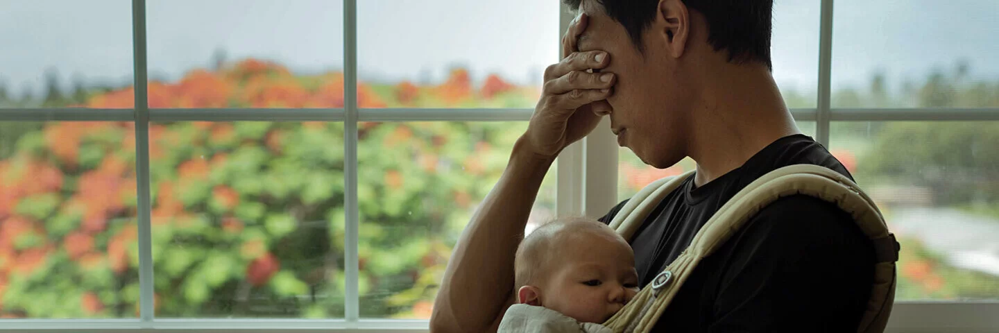 Ein Vater mit Baby vor der Brust reibt sich müde die Augen – er leidet unter Schlafmangel.