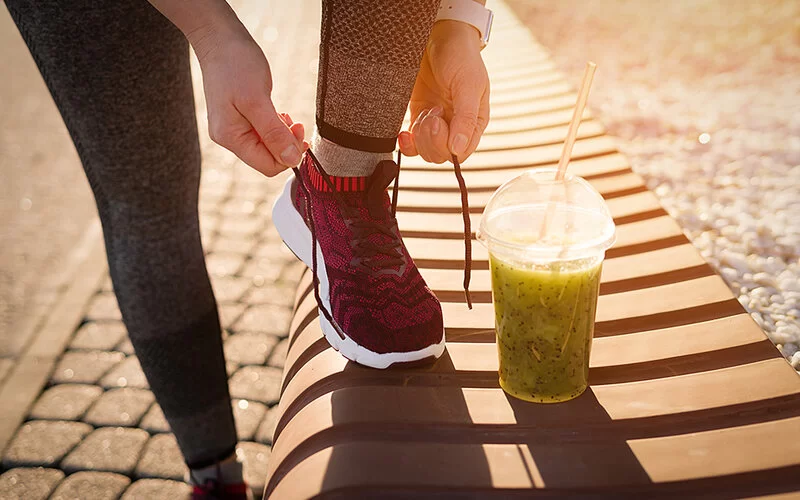 Eine Joggerin bindet sich die Laufschuhe zu, während vor ihr ein frischer Smoothie bereitsteht.