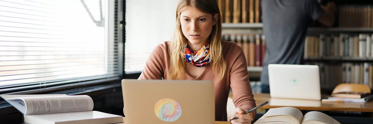 Eine junge Frau sitzt an einem Tisch und schaut auf einen Laptop. Auf dem Tisch liegen ein paar aufgeschlagene Bücher.