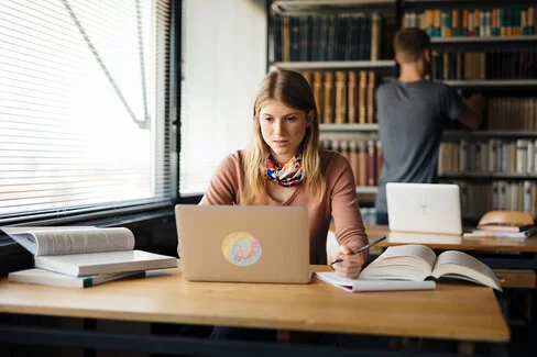 Eine junge Frau sitzt an einem Tisch und schaut auf einen Laptop. Auf dem Tisch liegen ein paar aufgeschlagene Bücher.