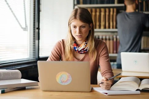 Eine junge Frau sitzt an einem Tisch und schaut auf einen Laptop. Auf dem Tisch liegen ein paar aufgeschlagene Bücher.