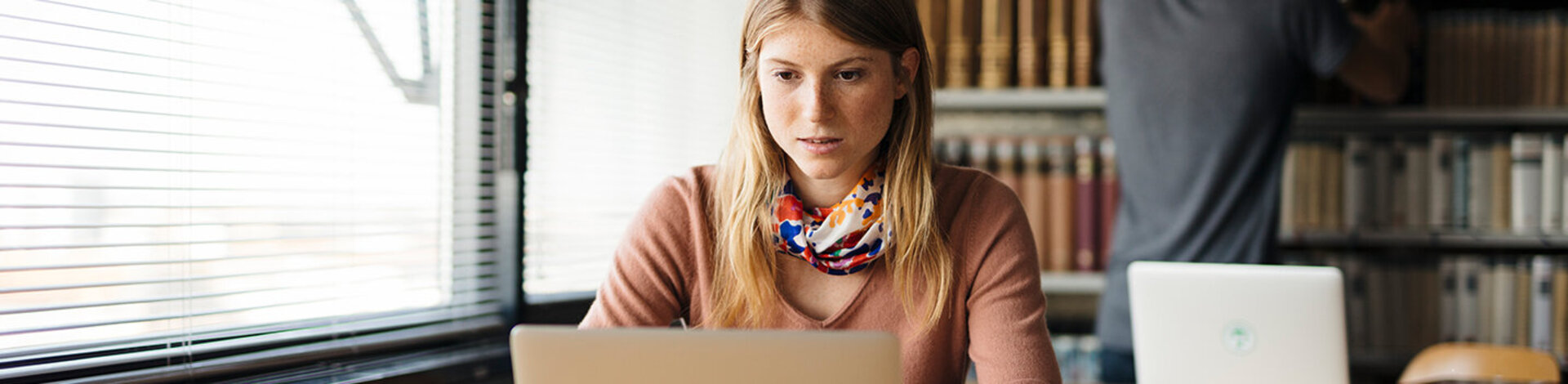 Eine junge Frau sitzt an einem Tisch und schaut auf einen Laptop. Auf dem Tisch liegen ein paar aufgeschlagene Bücher.