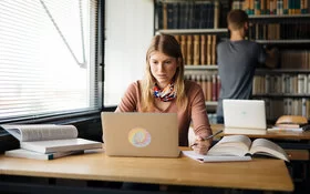 Eine junge Frau sitzt an einem Tisch und schaut auf einen Laptop. Auf dem Tisch liegen ein paar aufgeschlagene Bücher.