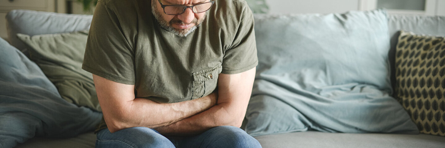Ein Mann sitzt in gebeugter Haltung auf einem Sofa und verschränkt die Arme haltend auf seinem Bauch.