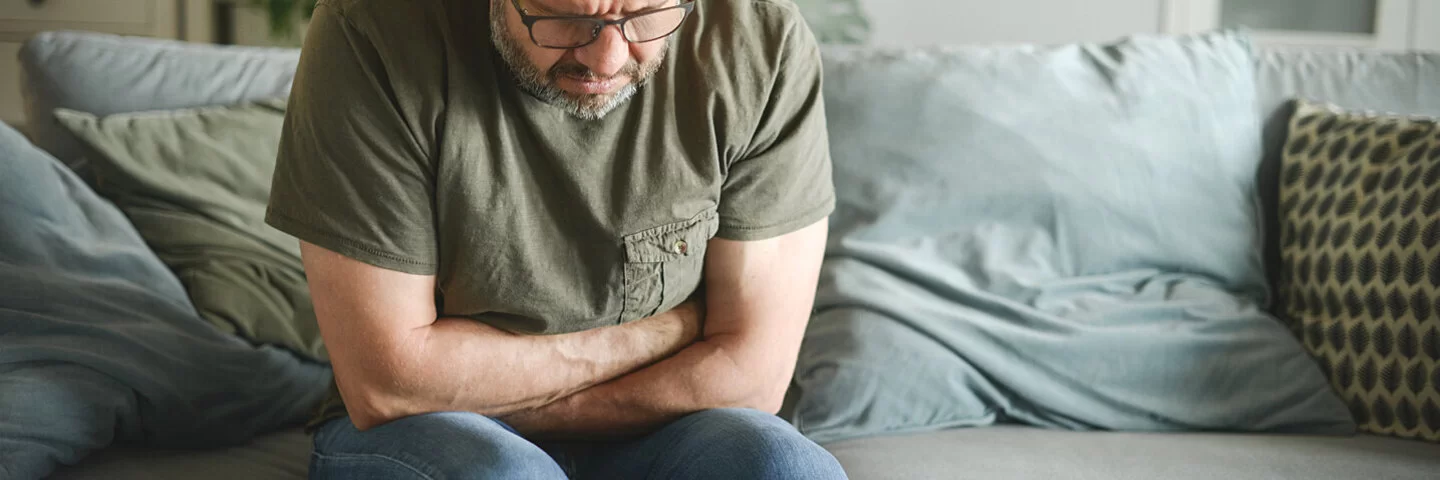 Ein Mann sitzt in gebeugter Haltung auf einem Sofa und verschränkt die Arme haltend auf seinem Bauch.