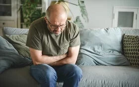 Ein Mann sitzt in gebeugter Haltung auf einem Sofa und verschränkt die Arme haltend auf seinem Bauch.