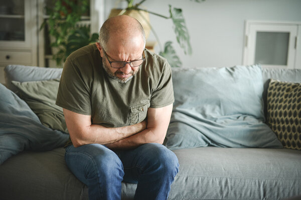 Ein Mann sitzt in gebeugter Haltung auf einem Sofa und verschränkt die Arme haltend auf seinem Bauch.