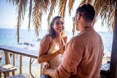 Eine blonde Frau und ein dunkelhaariger Mann stehen unter Palmen an einer Strandbar und unterhalten sich lächelnd, im Hintergrund das Meer.