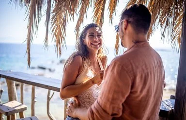 Eine blonde Frau und ein dunkelhaariger Mann stehen unter Palmen an einer Strandbar und unterhalten sich lächelnd, im Hintergrund das Meer.