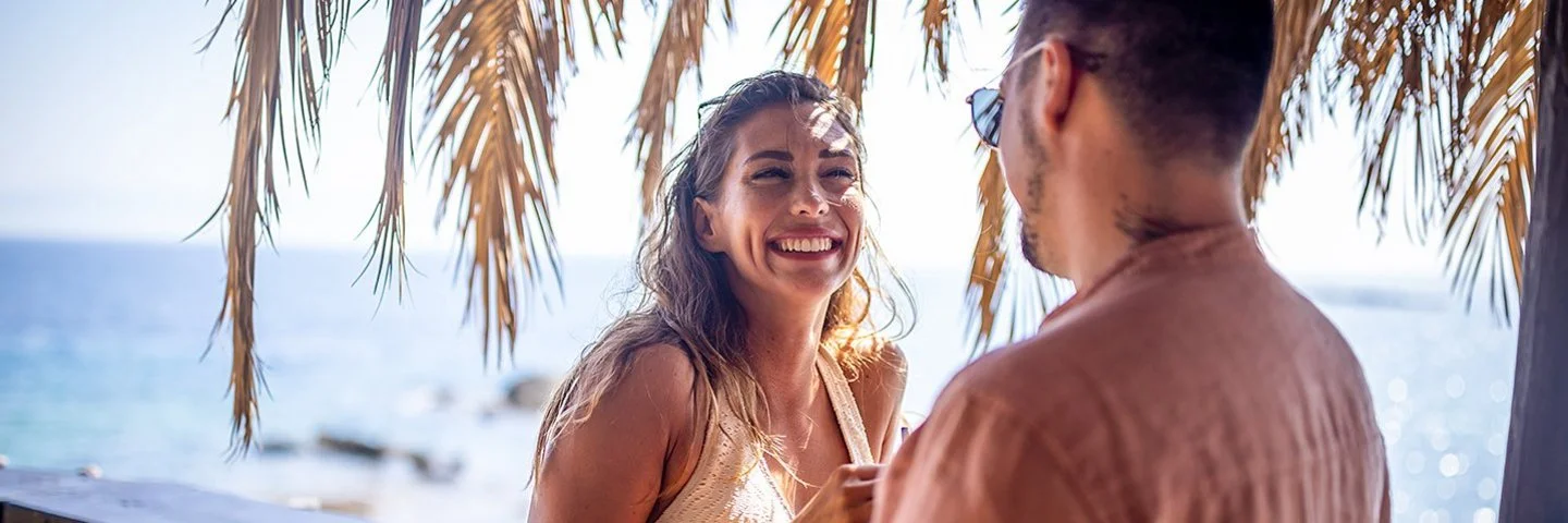 Eine blonde Frau und ein dunkelhaariger Mann stehen unter Palmen an einer Strandbar und unterhalten sich lächelnd, im Hintergrund das Meer.