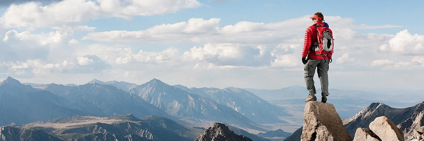 Ein Mann steht auf einer Felsspitze und blickt auf eine unter ihm liegende karge Gebirgslandschaft.